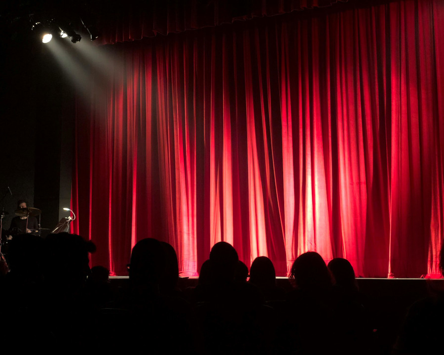 Tecnología para la Vida TXLV Torrelodones - MicroMagazine de FUNTESO, Fundación Tecnología Social imagen de un escenario en el que destaca una cortina roja de teatro, varias personas sentadas en sus butacas de espalda y varios focos, además de un equipo musical